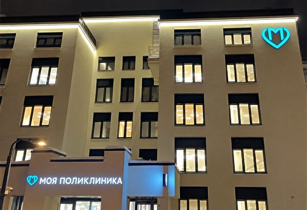 Фасад здания с освещёнными окнами, бирюзовой эмблемой и вывеской «Моя поликлиника» у входа.