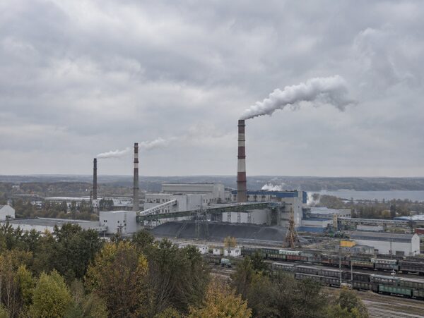 Панорама промышленных зданий и труб с тонкими шлейфами выбросов под серым небом.