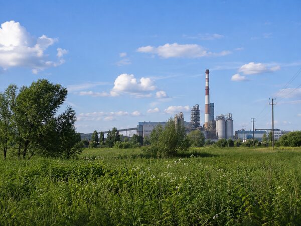 Зелёное поле и деревья на переднем плане, промышленные сооружения и трубы вдали.