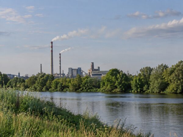 Водоём и зелёные деревья на переднем плане, промышленный комплекс с трубами вдали.