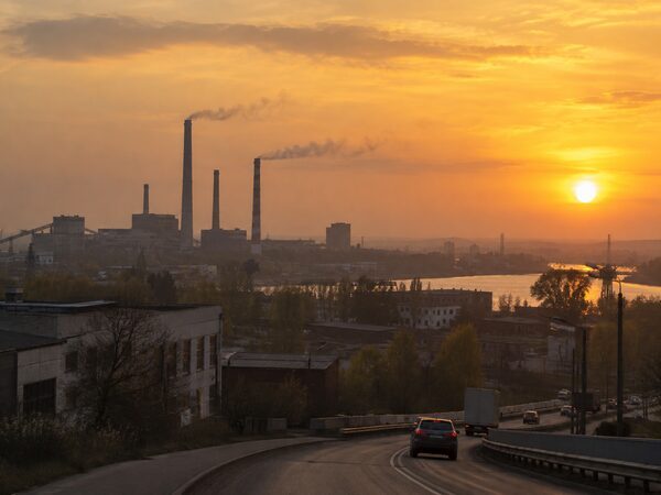 Промышленные здания и трубы видны в закатной дымке за дорогой с транспортом.