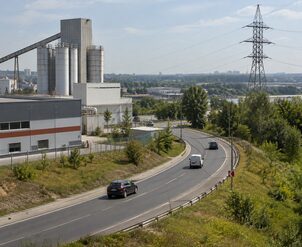 Изогнутая автомобильная дорога с транспортом проходит рядом с промышленными сооружениями и линией электропередачи.