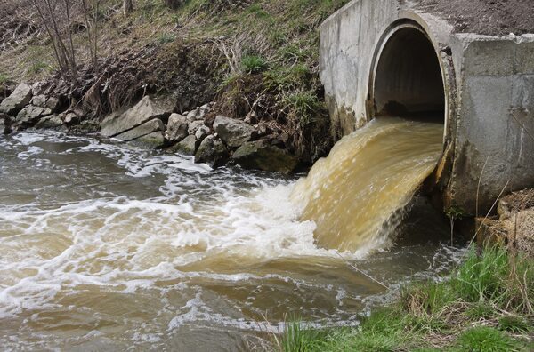 Мутная вода вытекает из бетонной трубы в открытый водоток с каменистым берегом.