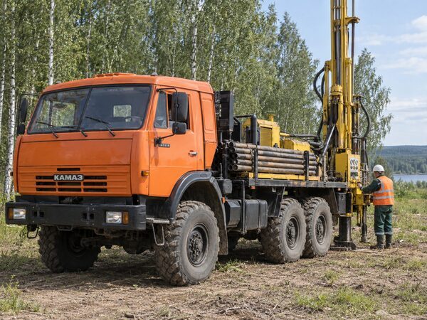 Оранжевый трёхосный КамАЗ с буровой мачтой и трубами; у пульта стоит работник в зелёной спецодежде, оранжевом жилете и белой каске.