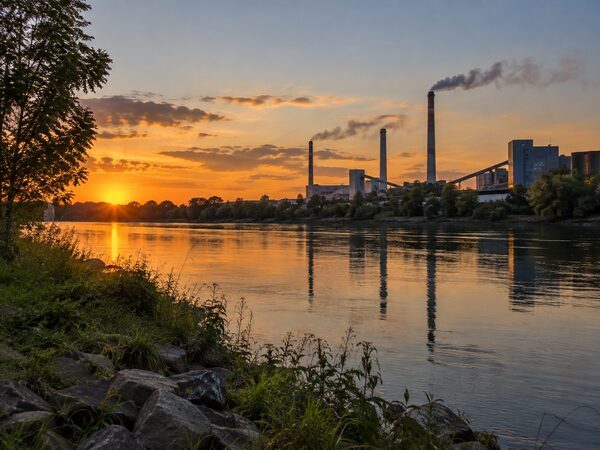 Промышленные трубы с дымовыми шлейфами отражаются в воде на фоне оранжевого закатного неба.